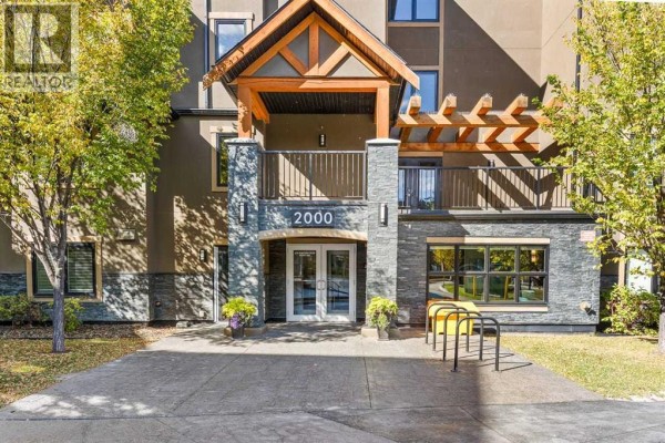 Apartment building entrance with double glass doors, stone accents, and wooden pergola. - 2405, 450 Kincora Glen Road Nw, Calgary