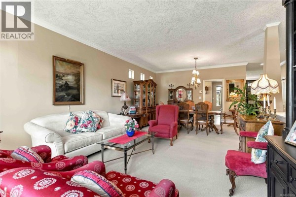 Living room with a sofa, armchair, coffee table, and fireplace, open to the kitchen. - 2409 Poplar Dr, Sooke
