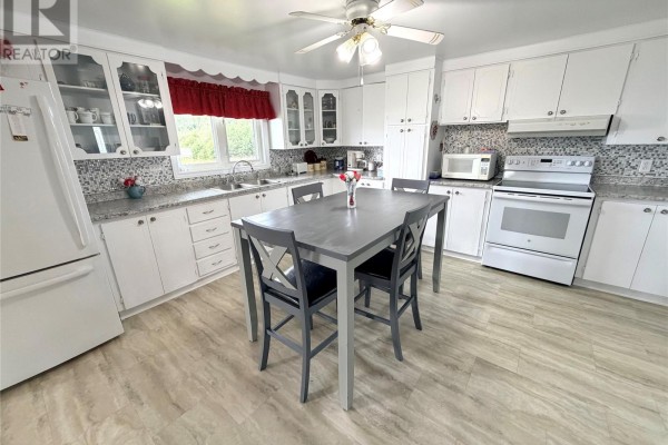 White kitchen cabinets, countertops, and appliances, with a dining table in the center. - 242 Main Street, Norris Arm North