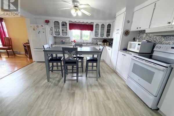 View of the kitchen with white cabinets and appliances, and a dining table. An opening leads to a room with wood floors. - 242 Main Street, Norris Arm North