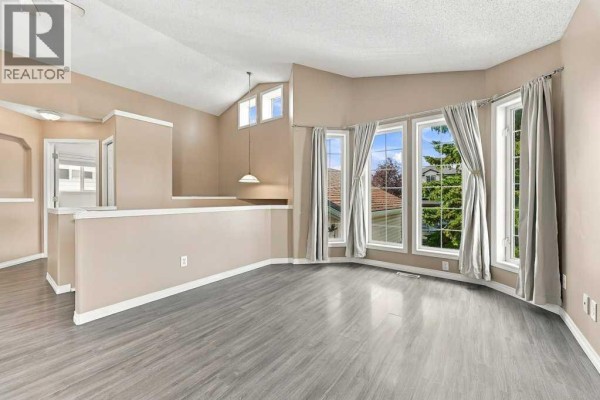 Living room with beige walls, vaulted ceiling, and large bay windows overlooking greenery. - 242 Springs Place Se, Airdrie