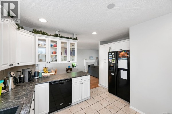 White kitchen cabinets, black dishwasher, black refrigerator, and a view into a living room. - 2437 Benko Rd, Mill Bay