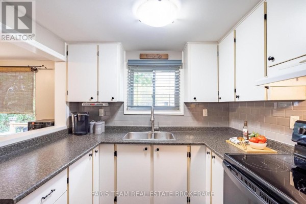 View of a U-shaped kitchen with white cabinets, dark countertops, a double sink, and a window. - 244/250 Dignard Avenue, Tay