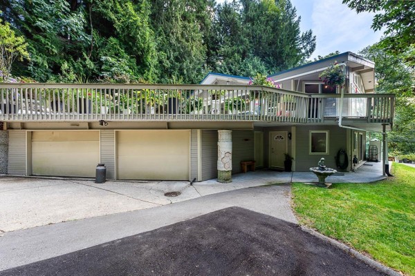 Close-up view of the three-car garage and front entrance of a grey house with a deck above. - 24400 56 Avenue, Langley