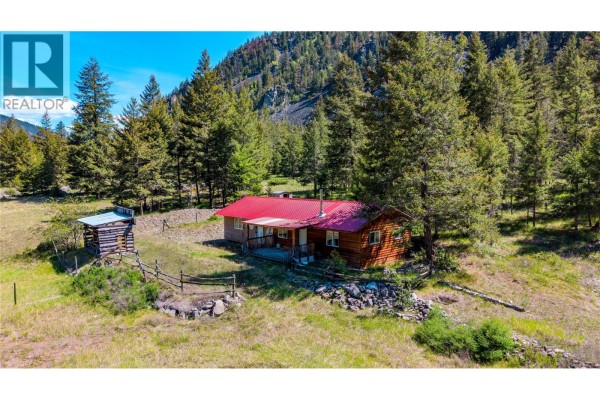 Log-sided house with a red metal roof, surrounded by trees and grassy terrain. - 2451 Hwy 3 E, Princeton
