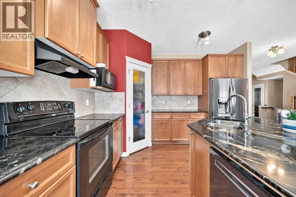 Close-up view of the kitchen's black stove, granite countertop, wood cabinets, and stainless steel refrigerator. - 246 Crystal Green Point, Okotoks