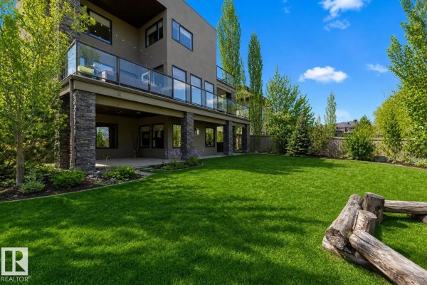 Backyard view of the house with a covered patio, green lawn, and mature trees surrounding the property. - 2464 Cameron Ravine Dr Nw, Edmonton