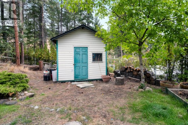 Close-up view of a small white shed with a teal door, surrounded by trees and stacked firewood. - 2466 Alexandria Way, West Kelowna