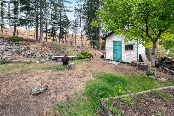 Backyard with a white shed, garden beds, a stone bench, and a treed landscape with a grassy area. - 2466 Alexandria Way, West Kelowna