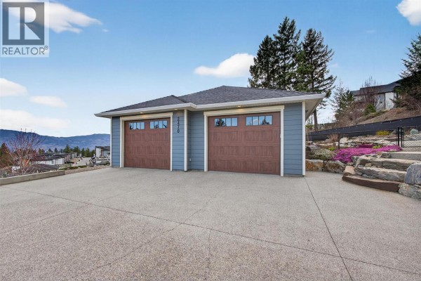 Attached two-car garage with brown wooden doors and gray siding, set against a backdrop of trees and sky. - 2478 Stone Grove Crescent, West Kelowna