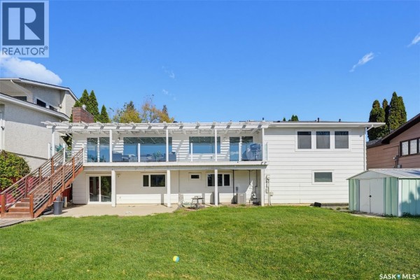 Rear exterior of a white house with a large deck and backyard, overlooking a grassy area. - 25 Capilano Drive, Saskatoon