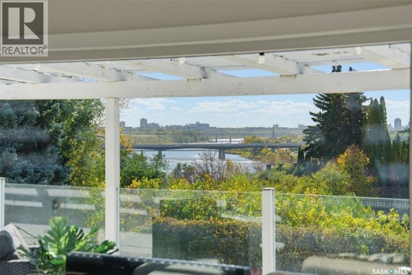 View of a river and city skyline through a glass-railed deck, with outdoor seating. - 25 Capilano Drive, Saskatoon