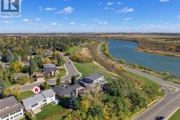 Aerial view of a residential street winding alongside a wide river with scattered trees. - 25 Capilano Drive, Saskatoon