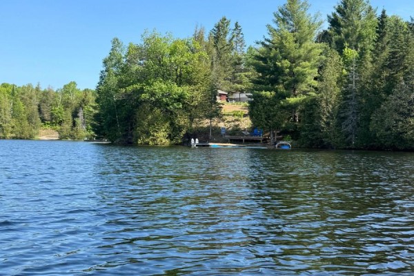 View of a wooden dock with chairs and a small building in the distance, set against a forested shoreline. - 25 Fire Route 330 Route, Trent Lakes