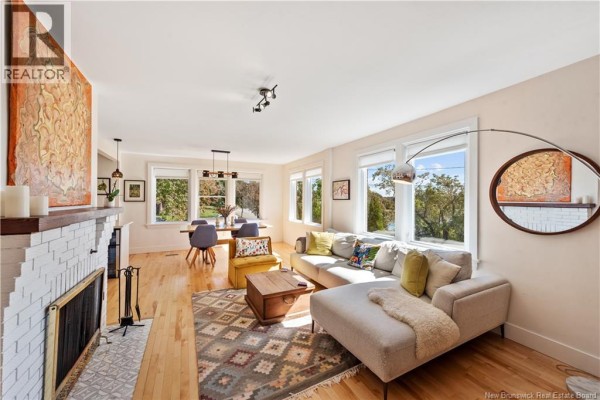 Living room with a fireplace, sectional sofa, coffee table, and large windows overlooking trees. - 25 Hillcrest Drive, Moncton