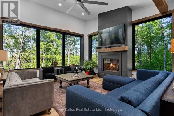 Living room with a large fireplace, a blue sectional sofa, and chairs, surrounded by expansive windows overlooking a forest. - 25 Natures Way, Huntsville