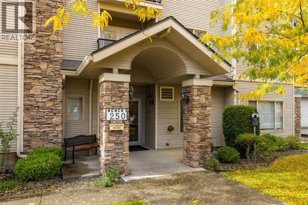 Entrance to building 250, Bench 1, with stone pillars and autumn trees. - 250 Dougall Road N Unit# 208, Kelowna
