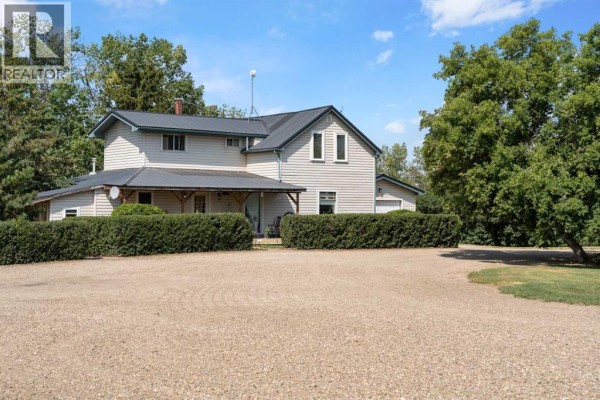 Two-story house with beige siding, a dark metal roof, and surrounding evergreen hedges. - 250035 Township Road 304, Rural Kneehill County