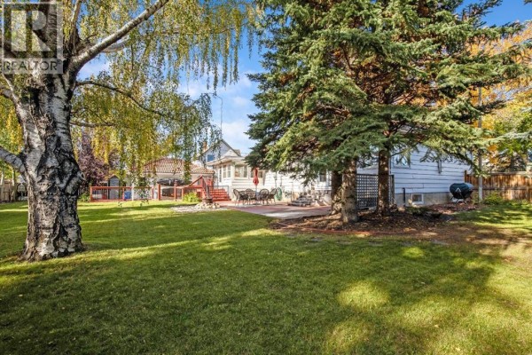 Backyard view with a large green lawn, mature trees, and a paved patio with outdoor furniture. - 2501 20 Street, Nanton