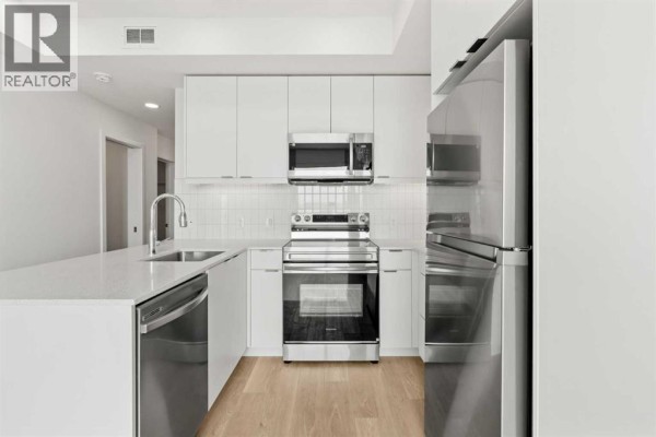 Close-up of a stainless steel range and refrigerator in a modern white kitchen with light wood flooring. - 2504, 8500 19 Avenue Se, Calgary