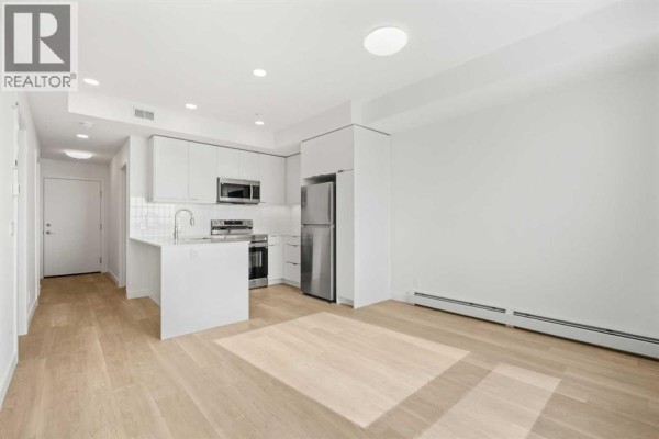 Open concept living area with a white kitchen, light wood floors, and white walls. - 2504, 8500 19 Avenue Se, Calgary