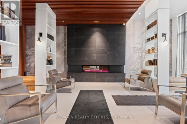 Apartment lobby lounge area with a modern black fireplace, grey chairs, white bookshelves, and beige seating. - 2505 - 297 Oak Walk Drive, Oakville