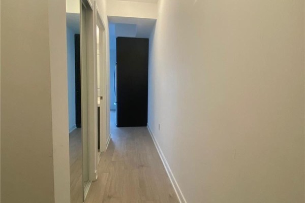 A hallway with light wood flooring, a mirrored closet door, and a black appliance. - 2506 - 8 Nahani Way, Mississauga
