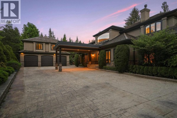 View of the triple car garage and covered entryway of a large home at dusk, with trees in the background. - 2514 East Road, Anmore