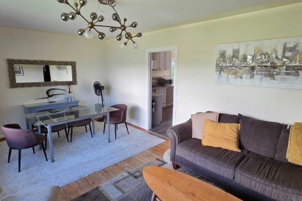 Living and dining area with a brown sofa, glass dining table, and dark chairs on a white rug. - 2515 Cochrane Road Nw, Calgary