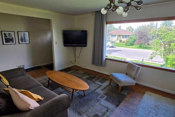 Living room with a sofa, coffee table, armchair, and a large picture window overlooking the street. - 2515 Cochrane Road Nw, Calgary
