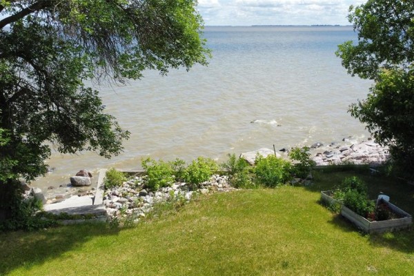 Waterfront view from a grassy yard with trees and rocky shoreline. - 253 Gimli Road, Whytewold
