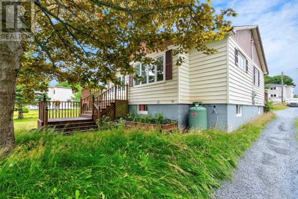 A light yellow house with vinyl siding, a wooden deck, and overgrown grass in the foreground. - 253 Water Street, Bay Roberts