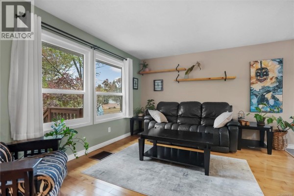 Living room with black leather sofa, wooden floors, and large windows with white curtains. - 2539 Cedar Hill Rd, Victoria