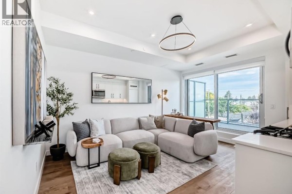 Living room with a sectional sofa, large mirror, and balcony access with city views. - 255, 110 18a Street Nw, Calgary