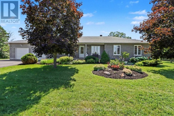 Front exterior of a grey brick house with a manicured lawn, flower beds, and mature trees. - 256 Orchard Crescent, Tay Valley