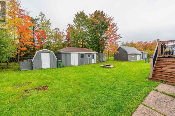 Backyard with green grass, three grey outbuildings, a fire pit, and a wooden deck with stairs. - 26 Poplar Drive, Lantz
