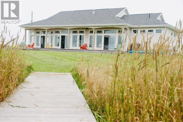 Wide shot of a grey house with a dark roof and large windows, set against a clear blue sky and grassy dunes. - 26 Roy's Lane, Augustine Cove