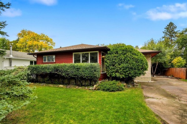 Front exterior of a red bungalow with dark brown roof, green shrubs, and driveway. - 26 Waterford Bay, Winnipeg