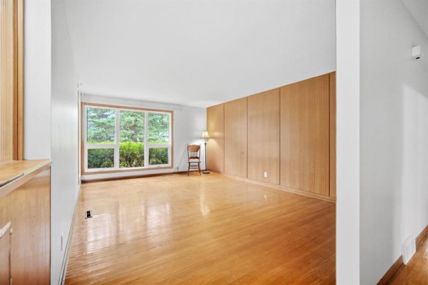 Empty living room with hardwood floors, large window overlooking greenery, and wood-paneled wall. - 26 Waterford Bay, Winnipeg