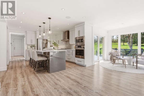 Open-concept kitchen and living area with white cabinets, a grey island, stainless steel appliances, and large windows. - 26 Weneil Drive, Hamilton