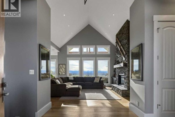 Living room with vaulted ceilings, large windows, and a stone-clad fireplace. - 260169 Mountain Ridge Place, Rural Rocky View County