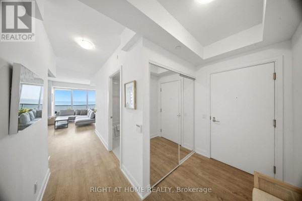 Apartment hallway with light wood floors, white walls, mirrored closet, and view of living room. - 2605 - 70 Annie Craig Drive, Toronto