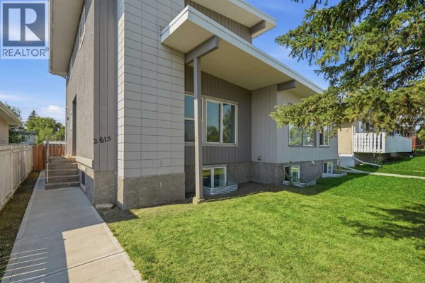Side exterior view of the multi-family home with gray siding and a concrete sidewalk. - 2615/2613 39 Street Se, Calgary