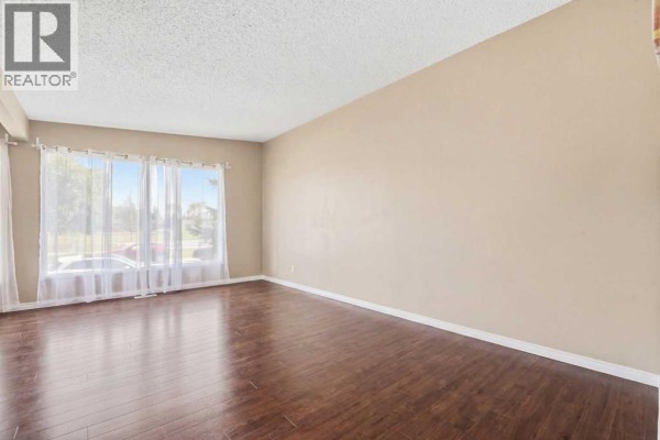 Empty living room with dark wood floors and large windows covered by sheer curtains. - 2615/2613 39 Street Se, Calgary