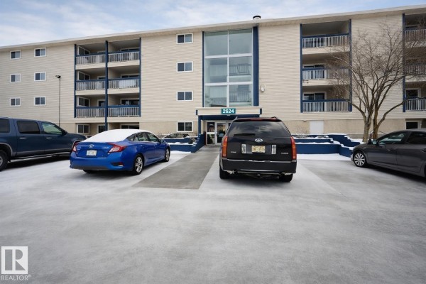 Exterior view of a beige apartment building with multiple balconies and a parking lot with snow. - 107, 2624 Millwoods Road East Nw, Edmonton
