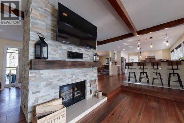 Stone fireplace with wood mantel, flat-screen TV, and a wooden crate with firewood. Open concept to kitchen. - 2631 Johnson Road, Fraser Lake