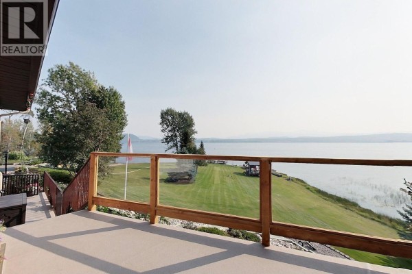 Deck with glass railing overlooking a green lawn, lake, and distant mountains under a hazy sky. - 2631 Johnson Road, Fraser Lake