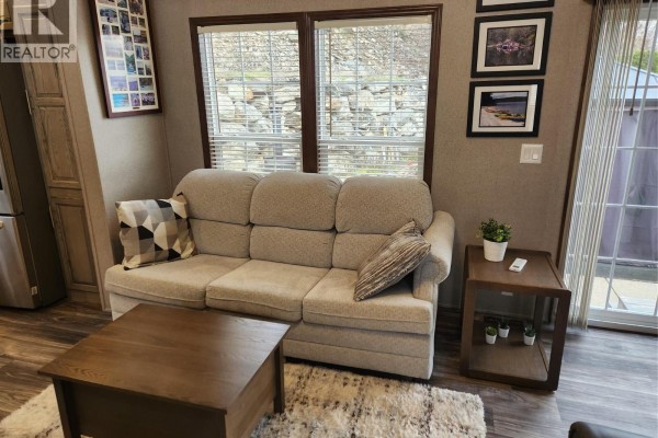 Living room with a beige sofa, coffee table, and windows overlooking a rocky hillside. - 2633 Squilax-anglemont Road Unit# 83, Lee Creek