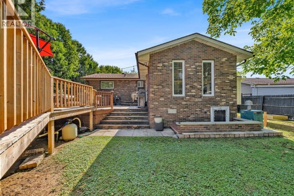 Backyard view of a brick house with a wooden deck, steps, a small patio, and green grass. - 2653 Concession Rd 12, Tecumseh