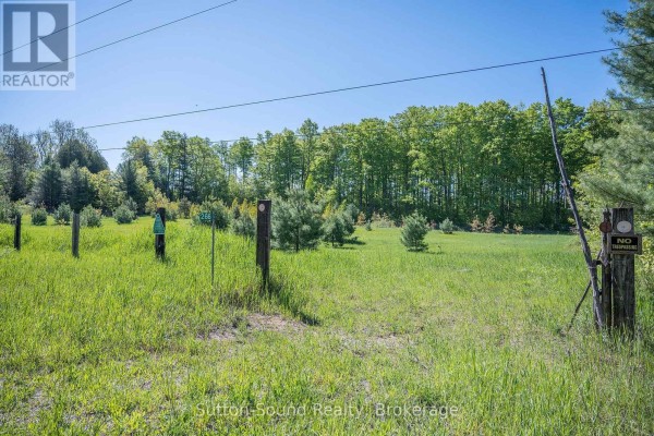View of a grassy field with wooden posts and signs, surrounded by young pine trees and a backdrop of green forest. - 266 Rankin Bridge Road, South Bruce Peninsula